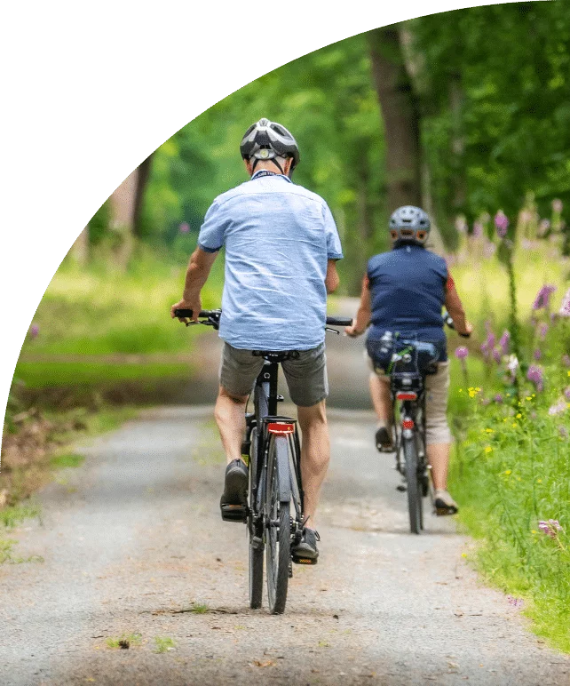 Two cyclists in helmets riding along a dirt path lined with trees and wildflowers