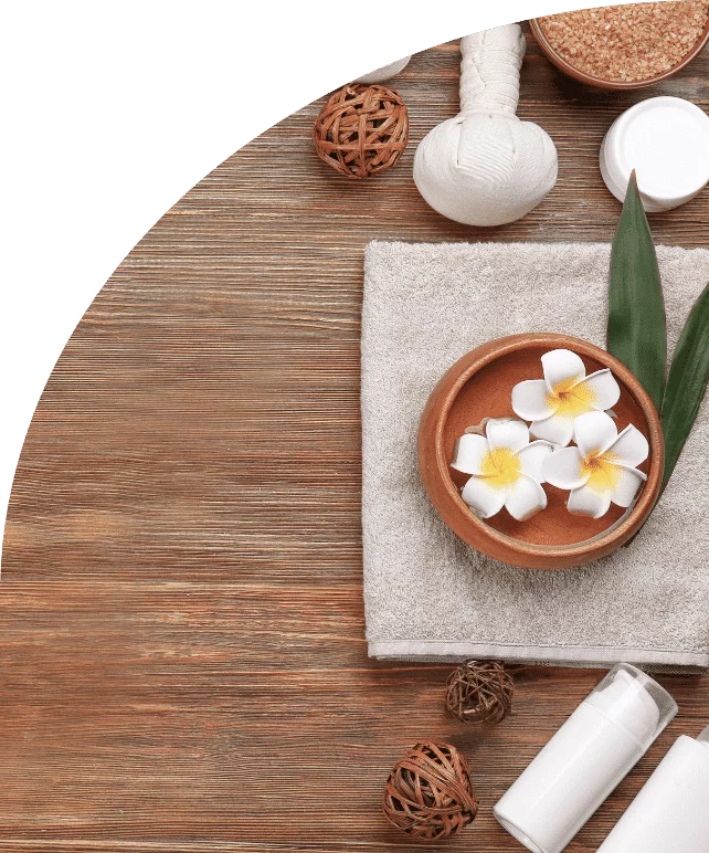 Serviette pliée, fleurs de frangipanier dans un bol, pochons de massage et flacons de soin sur une table en bois